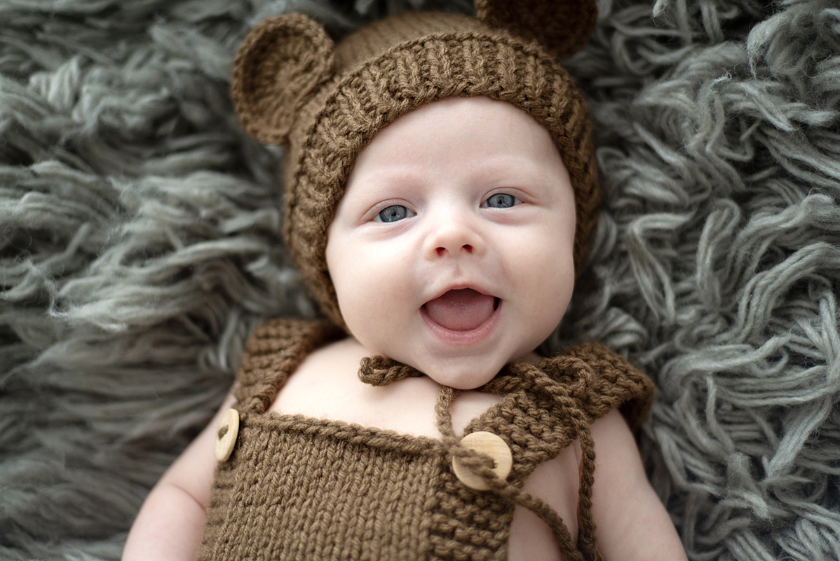 Bebê no ensaio de 2 meses usando touca de ursinho marrom sorri para a càmera