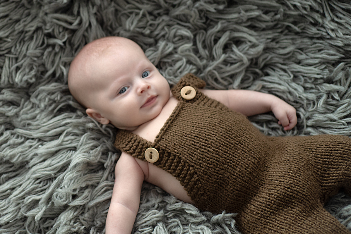 Bebê de 2 meses deitado no flokati cinza usando roupa de tricô marrom