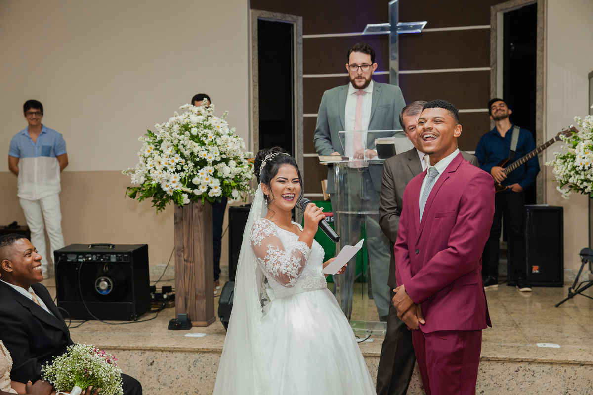 Noiva sorrindo com igreja decorada com flores brancas durante a leitura dos seus votos ao noivo, durante casamento em igreja evangélica em Pádua. RJ