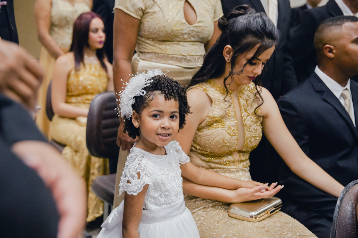 Daminha com tiara linda de flor na cabeça sorrindo perto de madrinha de vestido dourado durante casamento em igreja evangélica em Pádua. RJ