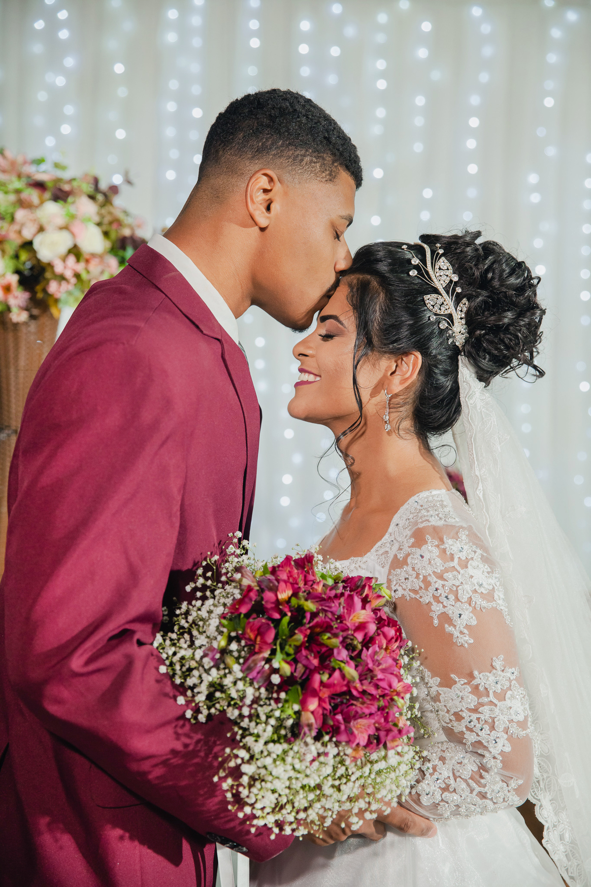 Noivo de terno vinho beijando a testa da noiva noiva com buque de flores rosa durante a festa de casamento em Santo Antônio de Pádua no rio de Janeiro.