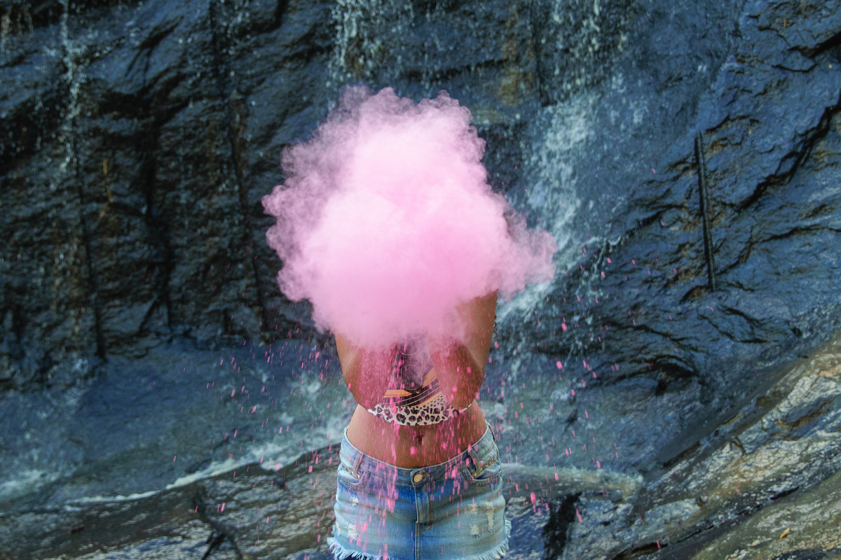 Aniversariante de 15 Anos com mini saia soprando pó colorido na cor rosa em cachoeira no seu ensaio fotográfico em uma fazenda em Santo Antônio de Pádua no interior do Rio de Janeiro.