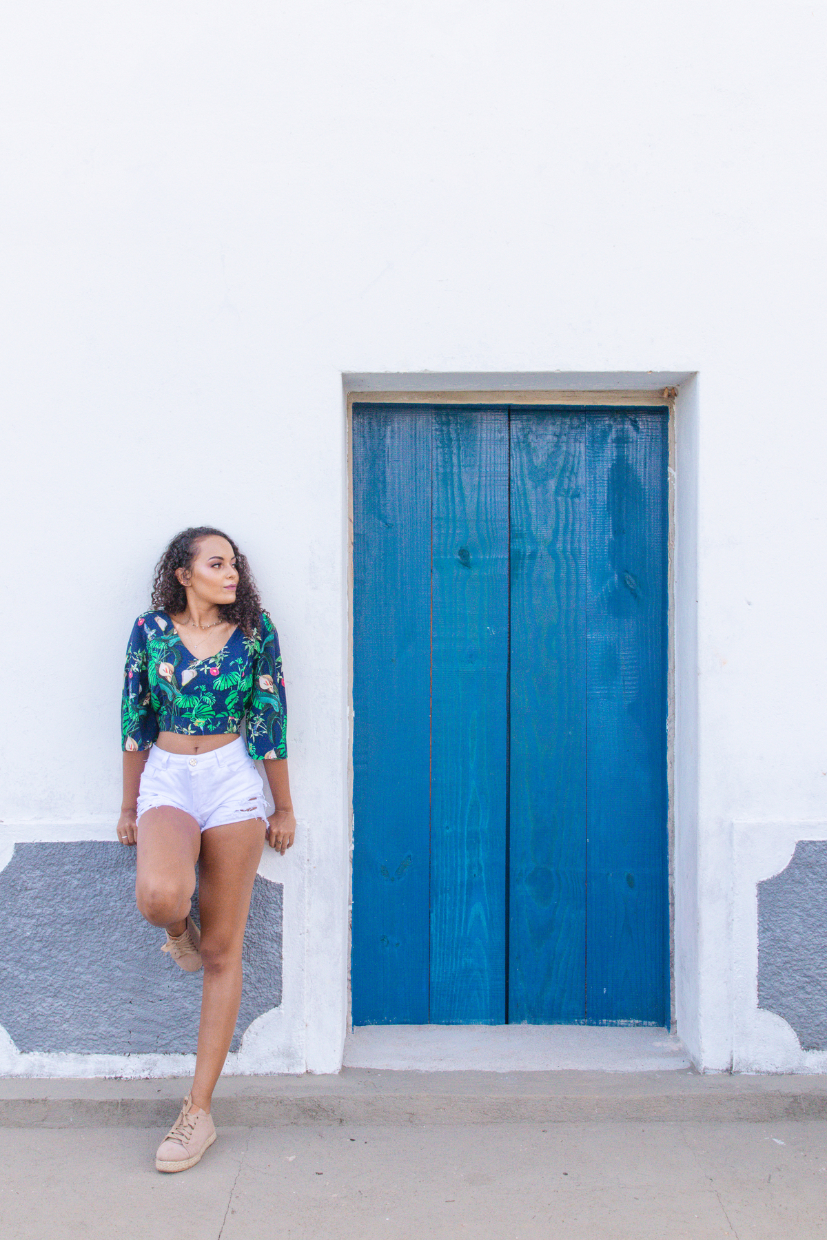 Menina de 15 anos com shorts branco e blusa estampada com tênis fazendo seu ensaio ao lado de uma porta azul em uma fazenda antiga em Santo Antônio de Pádua no interior do Rio de Janeiro.