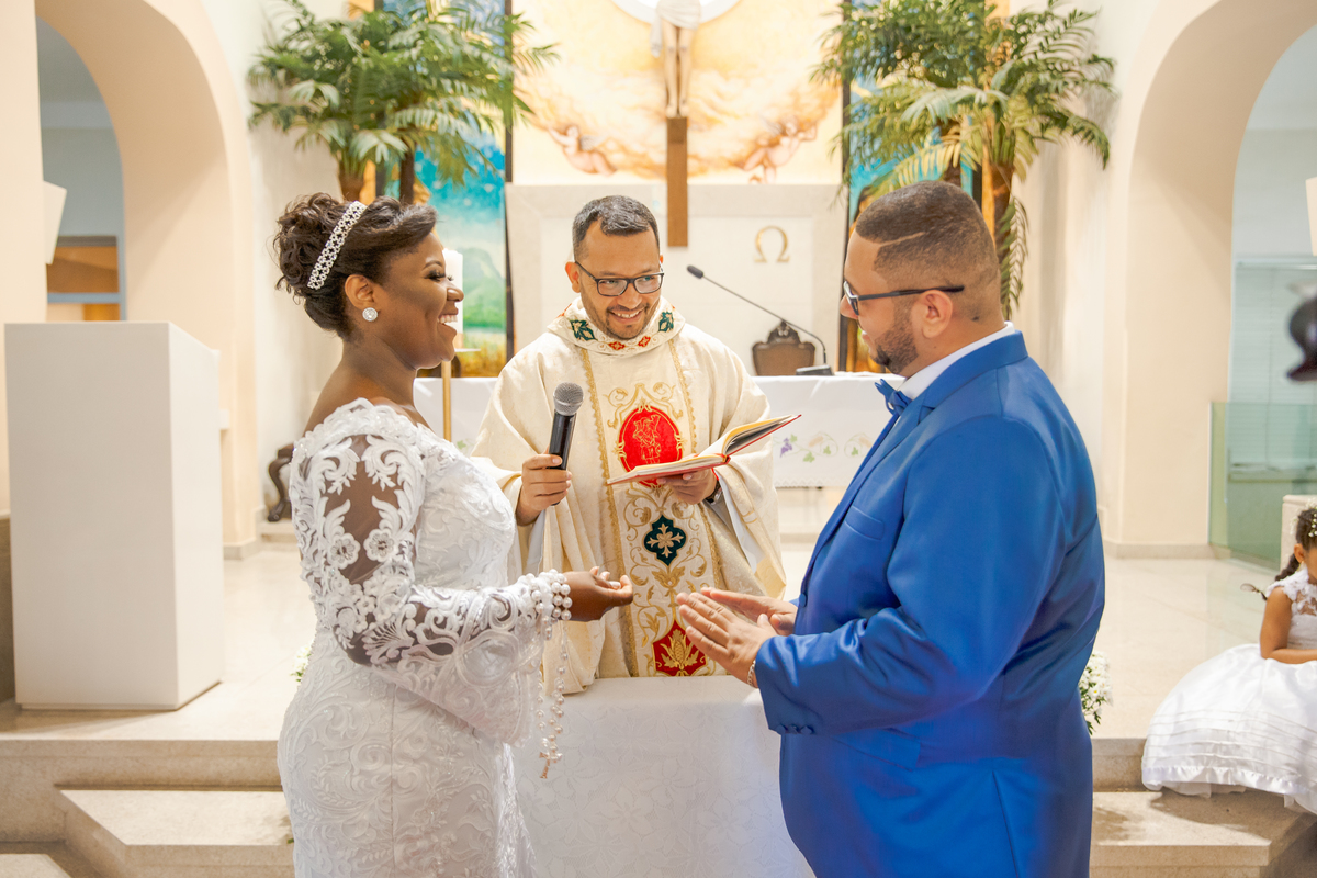 Noivos e padre sorrindo durante cerimônia  Paróquia de São José  de Leonissa em Itaocara no Rio de Janeiro. Noiva de vestido de renda lindo e noivo de terno azul de gravata borboleta.
