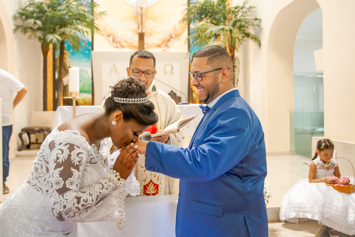 Noiva beijando a mão do noivo na cerimônia das alianças durante casamento na Paróquia de São José de Leonissa em Itaocara no Rio de Janeiro. Noiva de vestido de renda lindo e noivo de terno azul de gravata borboleta.