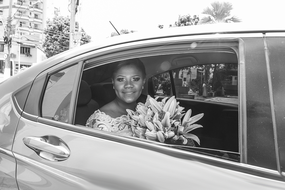 Foto em preta e branca de noiva saindo do carro, linda, maquiada com tiara de pedras nos cabelos e buque nas mãos  para entrar em se casamento na Paróquia de São José  de Leonissa em Itaocara no Rio de Janeiro.