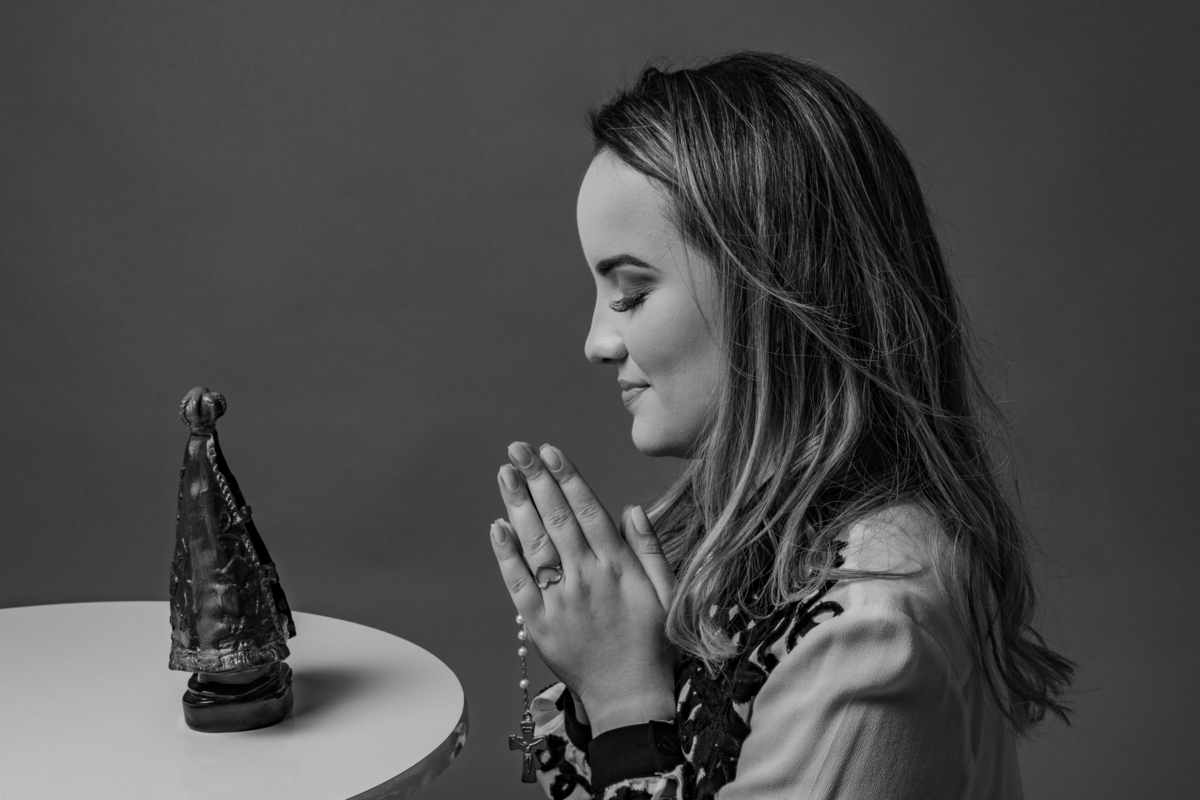 Formanda em psicologia linda com uma camisa branca agradecendo a uma imagem de nossa Senhora em ensaio realizado em estúdio com foto em Preto e Branca em Pádua no Rio de Janeiro