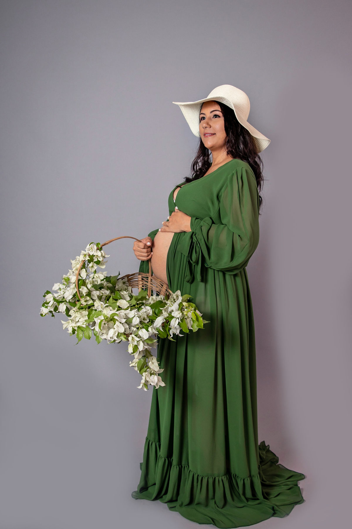 Grávida linda em seu ensaio de estúdio com fundo fotográfico cinza, com vestido longo verde com abertura na barriga segurando uma cesta de flores brancas e chapéu branco na cabeça em estúdio de Santo Antônio de Pádua RJ.
