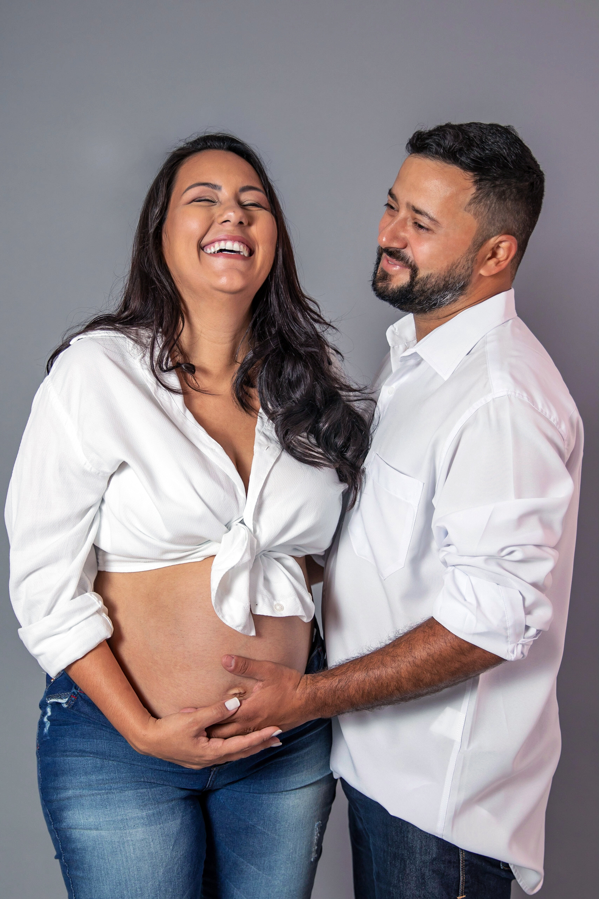 Ensaio gestante com calça jeans rasgada e camisa branca amarrada na barriga e salto alto branco. Ensaio feito em estúdio com fundo cinza em Santo Antônio de Pádua RJ.