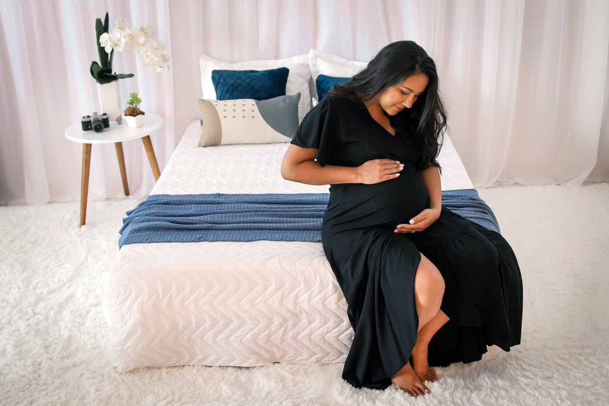 Linda gestante com vestido longo preto com a mão na barriga em estúdio com cenário de cama de casal em Pádua interior do Rio de Janeiro.