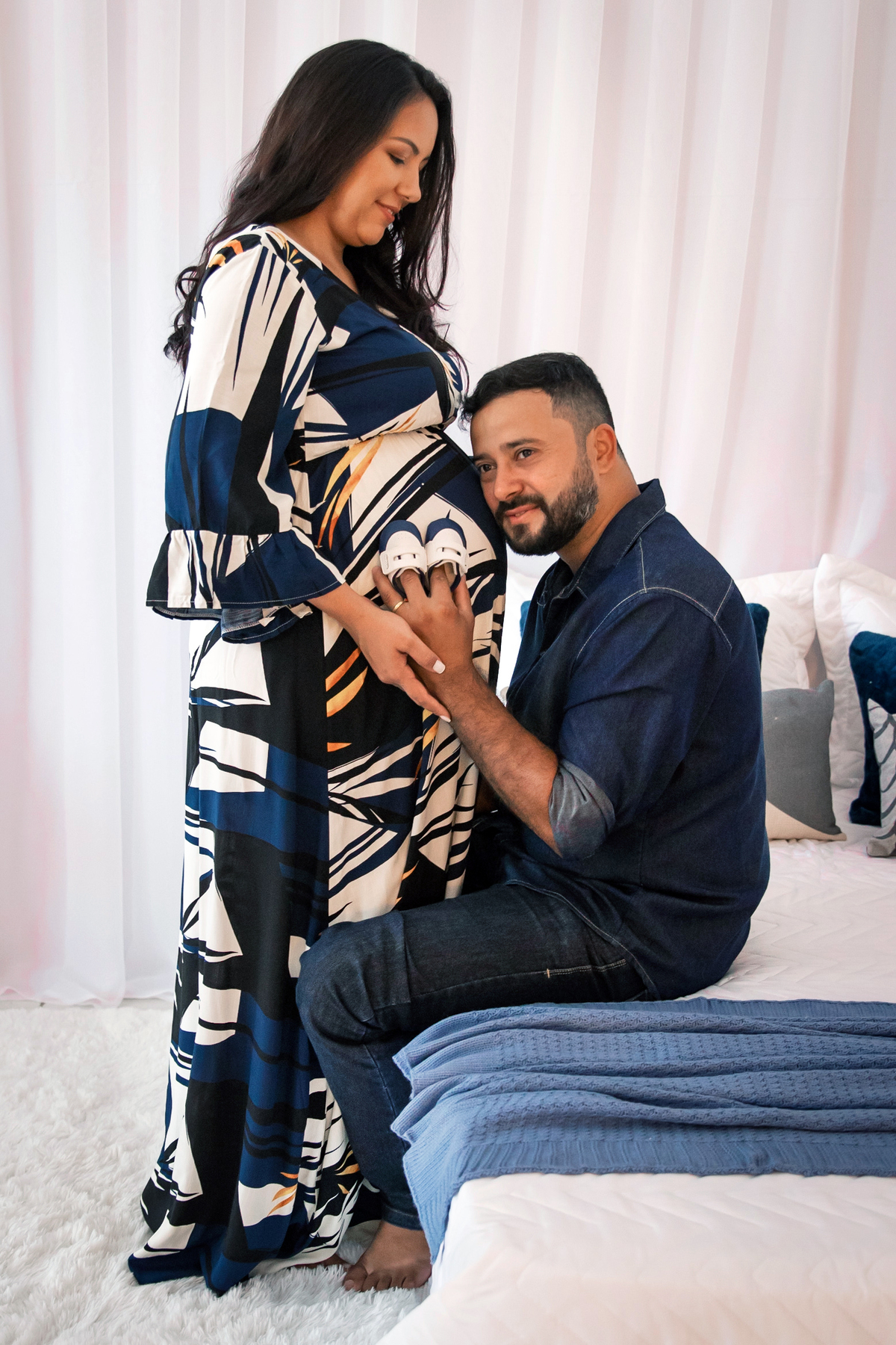 Grávida linda com vestido estampado azul longo e seu esposo fazendo carinho na barrida segurando sapatinho de bebe em quarto lindo montado em estúdio em Santo Antônio de Pádua RJ.