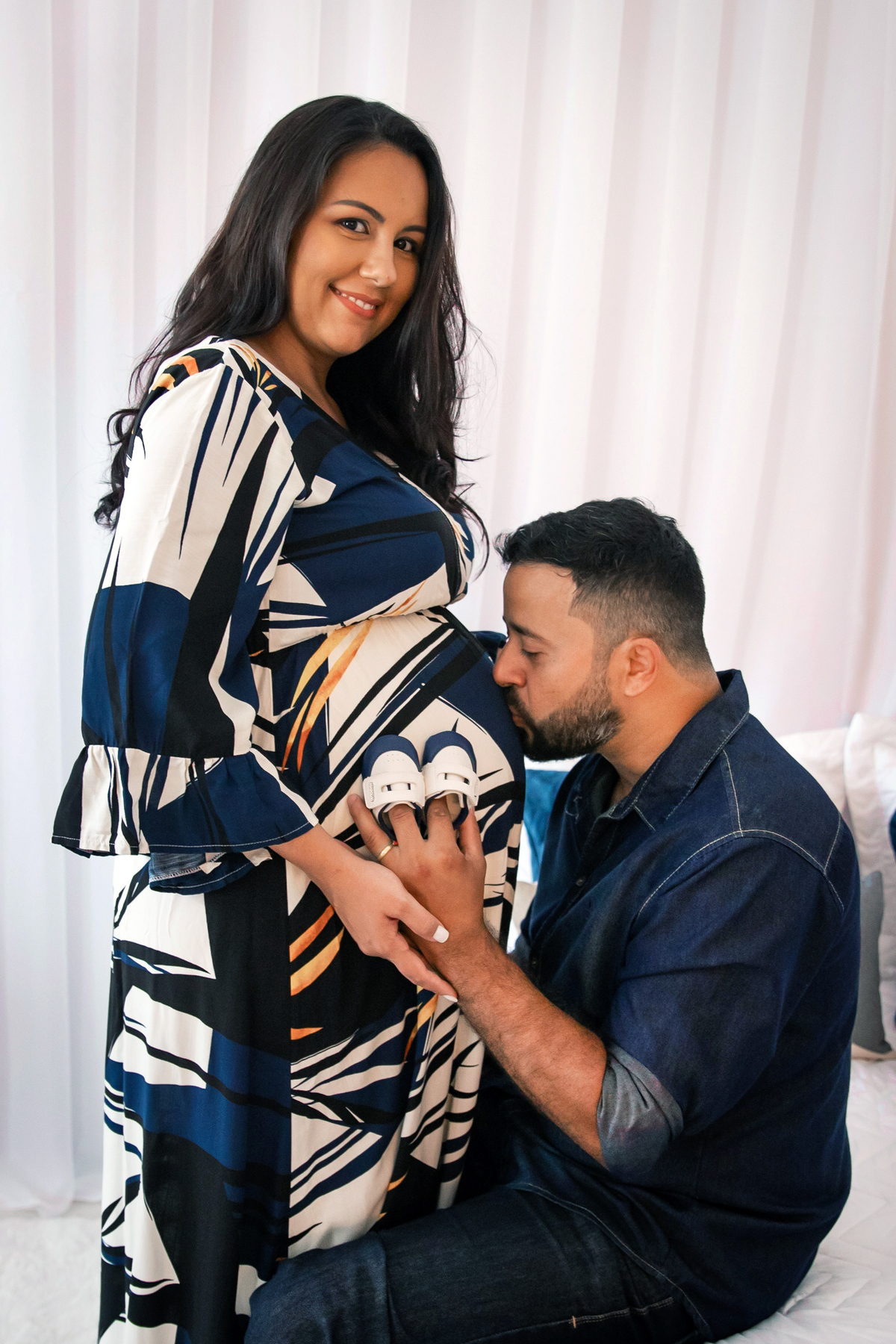 Grávida linda com vestido estampado azul longo e seu esposo fazendo carinho na barrida segurando sapatinho de bebe em quarto lindo montado em estúdio em Santo Antônio de Pádua RJ.estúdio em Santo Antônio de Pádua RJ.