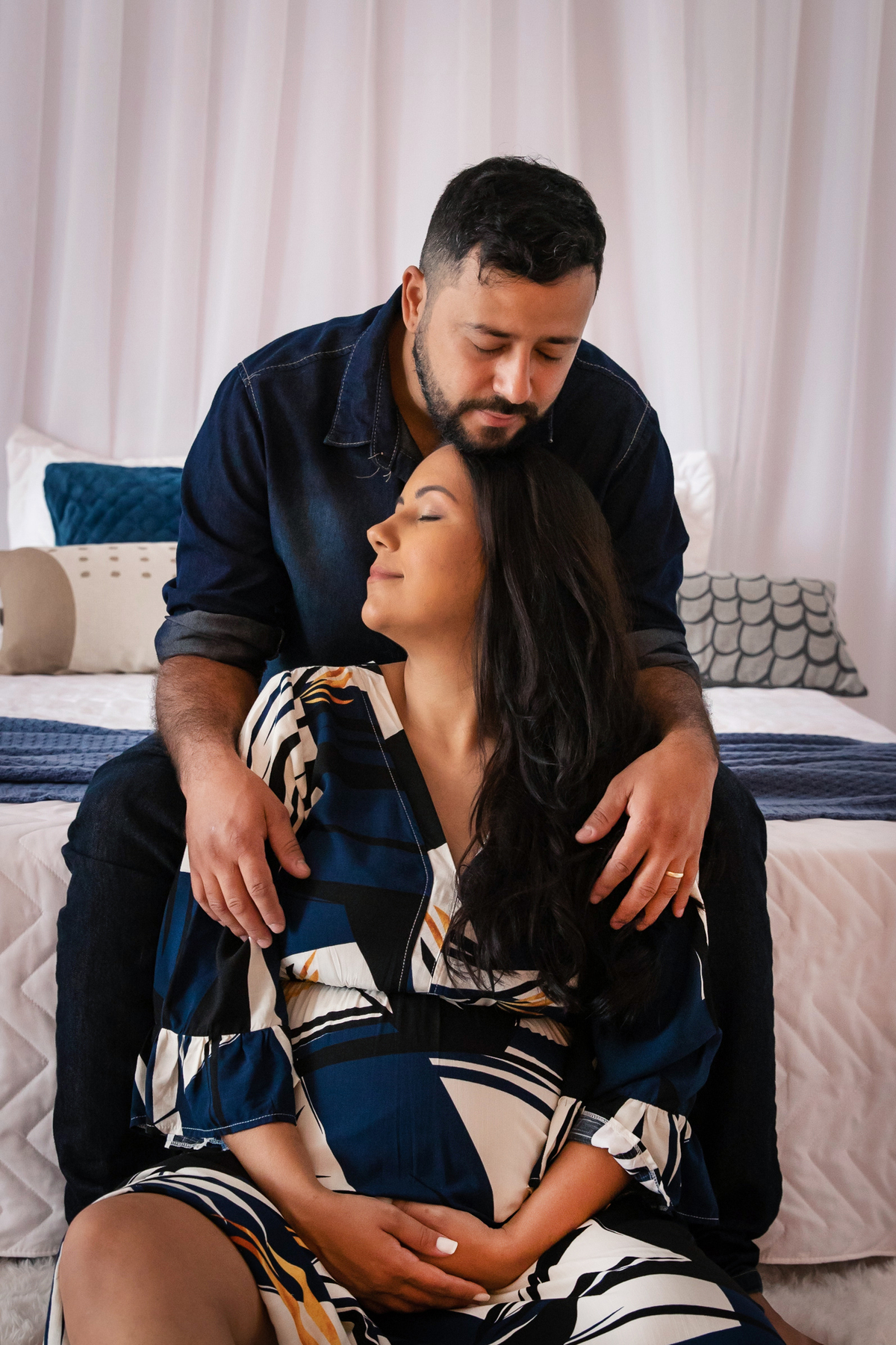 Grávida linda com vestido estampado azul longo e seu esposo fazendo carinho na barrida segurando sapatinho de bebe em quarto lindo montado em estúdio em Santo Antônio de Pádua RJ.