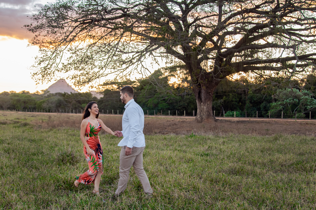  Casal abraçados em baixo de uma arvore enorme e linda em seu ensaio pre casamento. A noiva usa vestido laranja combinando como por do sol em Itaocara no interior do Rio de Janeiro.
