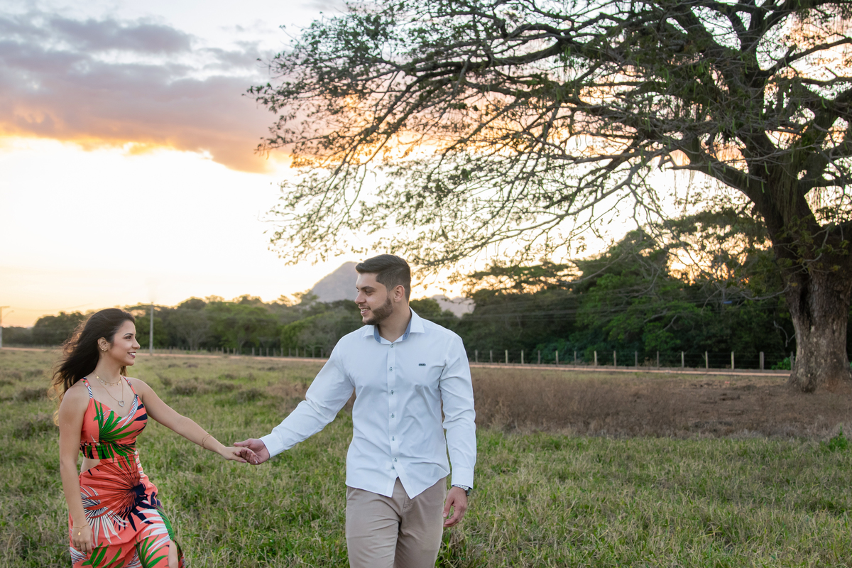  Casal caminhando em baixo de uma arvore enorme e linda em seu ensaio pre casamento. A noiva usa vestido laranja combinando como por do sol em Itaocara no interior do Rio de Janeiro.