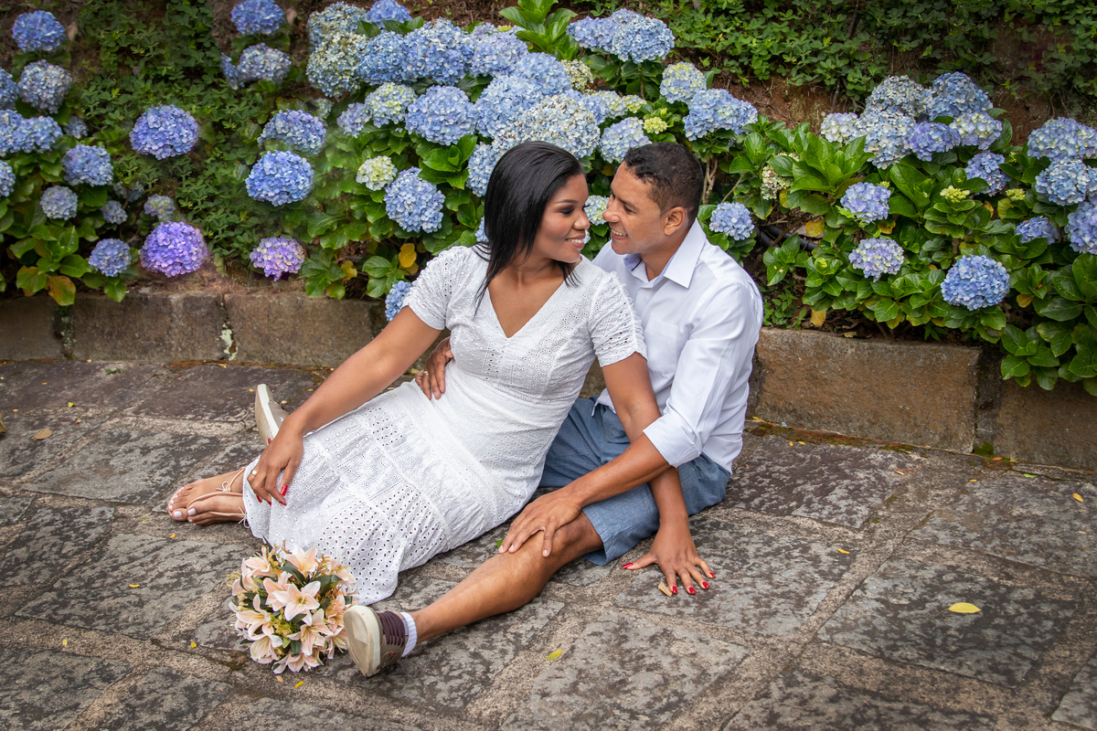 Noivos sentados para seu ensaio pré casamento em meio as hortênsias em dia nublado na serra de Teresópolis, noiva e noivo com roupas brancas em uma casa linda com muito verde e muitas flores e hortênsias azuis em Pàdua RJ.