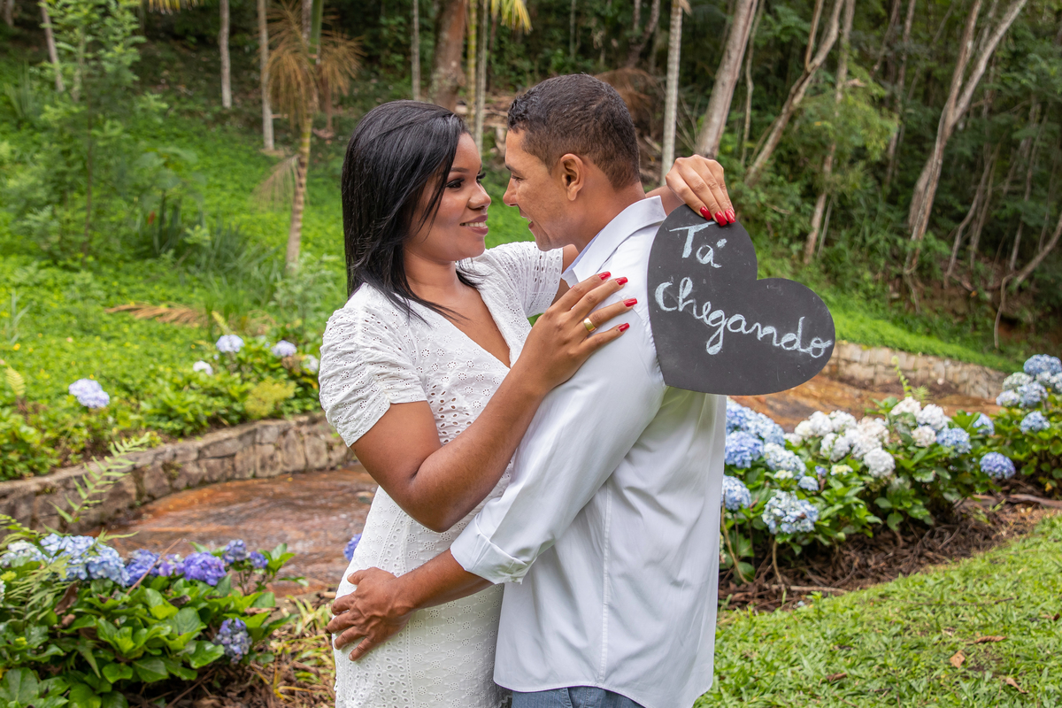 Noiva segurando um plaquinha escrito tá chegando em seu ensaio pre casamento na serra de Teresópolis, noiva e noivo com roupas brancas em uma casa linda com muito verde e muitas flores e hortênsias azuis em Pàdua RJ.
