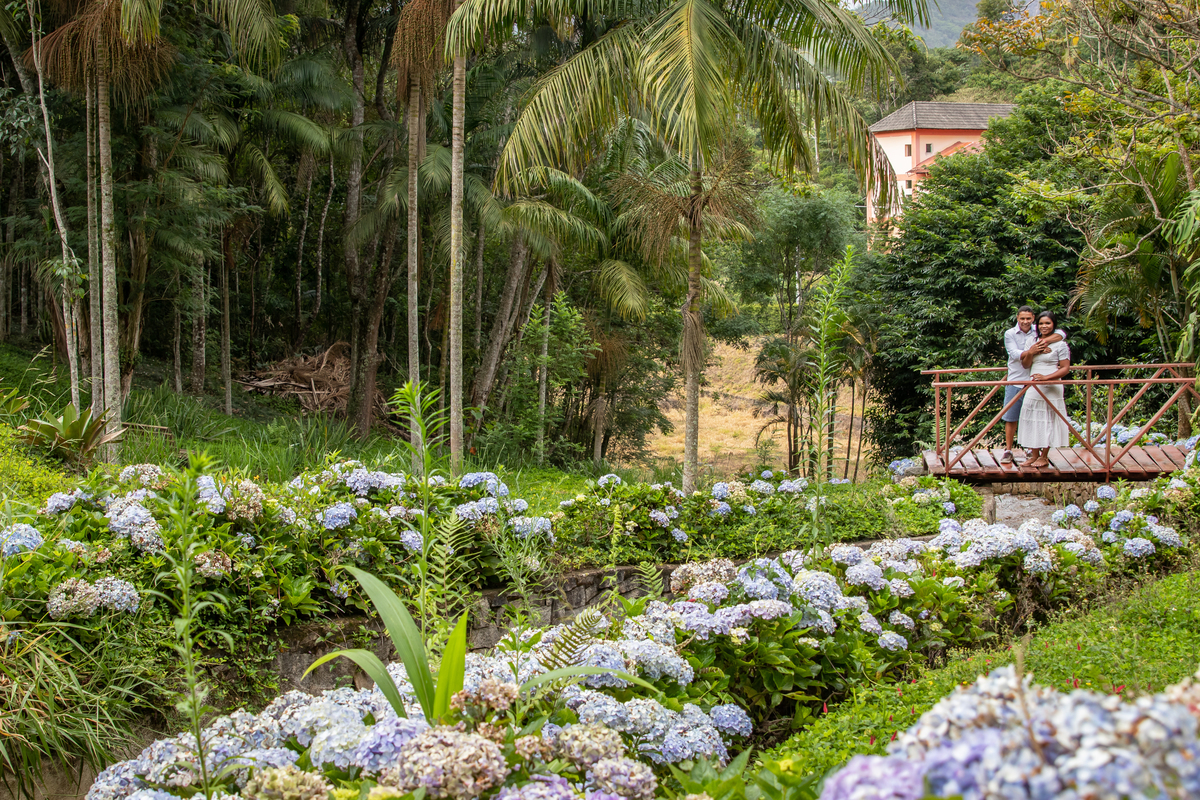 Noivos e hortênsias azuis em dia nublado na serra de Teresópolis, durante ensaio pré wedding noiva e noivo com roupas brancas em uma casa linda com muito verde e muitas flores e hortênsias azuis em Pàdua RJ.