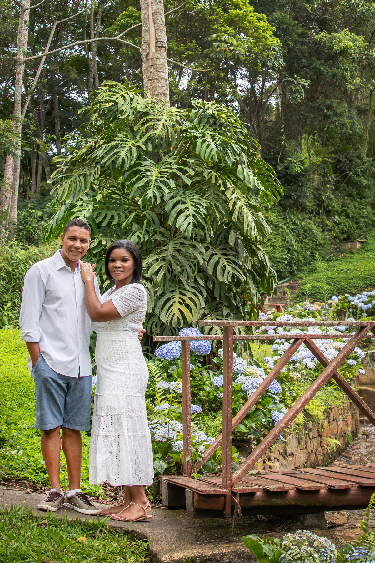 Noivos sorrindo durante ensaio fotográfico pré casamento na serra de Teresópolis, noiva e noivo com roupas brancas em uma casa linda com muito verde e muitas flores e hortênsias azuis em Pàdua RJ.