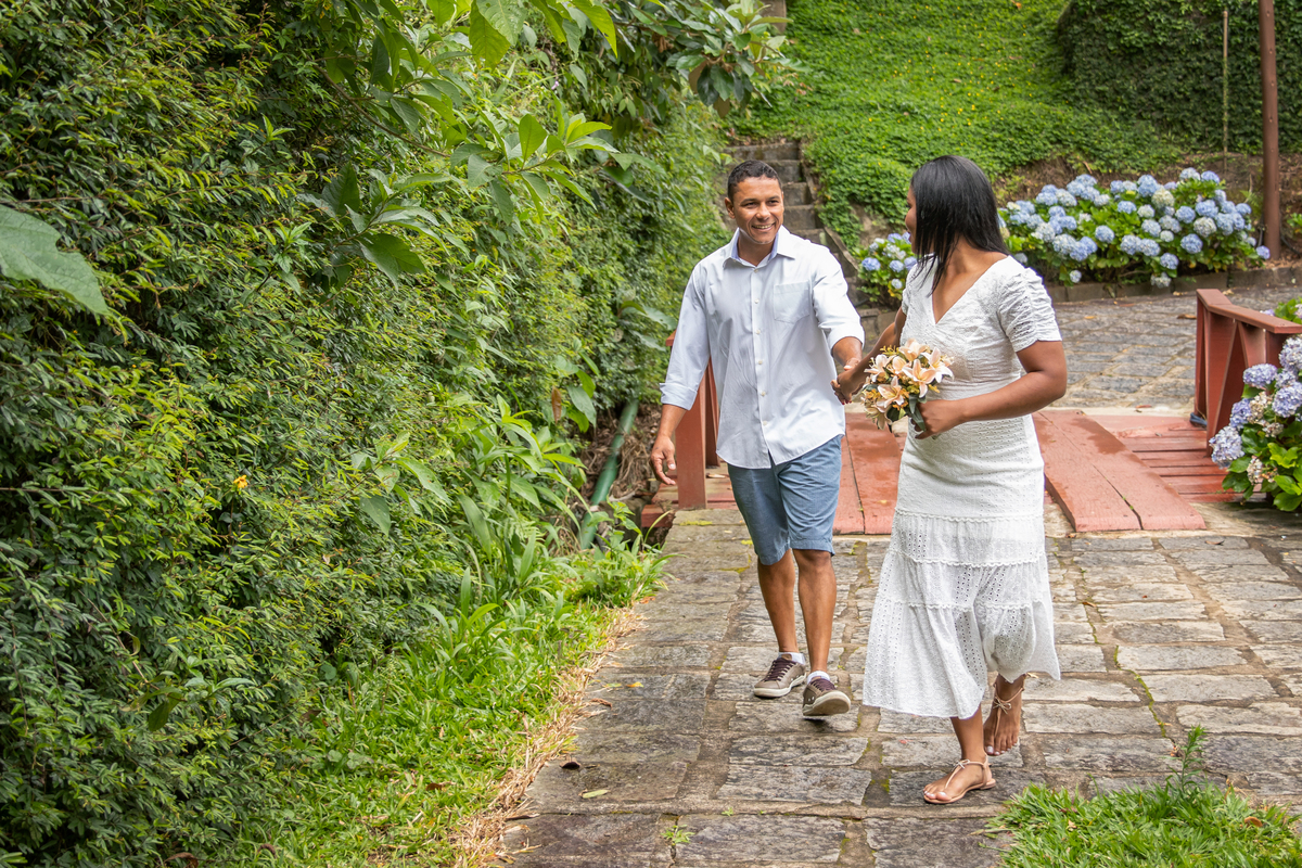 Noiva puxando o noivo feliz durante ensaio de casal antes do casamento na serra de Teresópolis, noiva e noivo com roupas brancas em uma casa linda com muito verde e muitas flores e hortênsias azuis em Pàdua RJ.