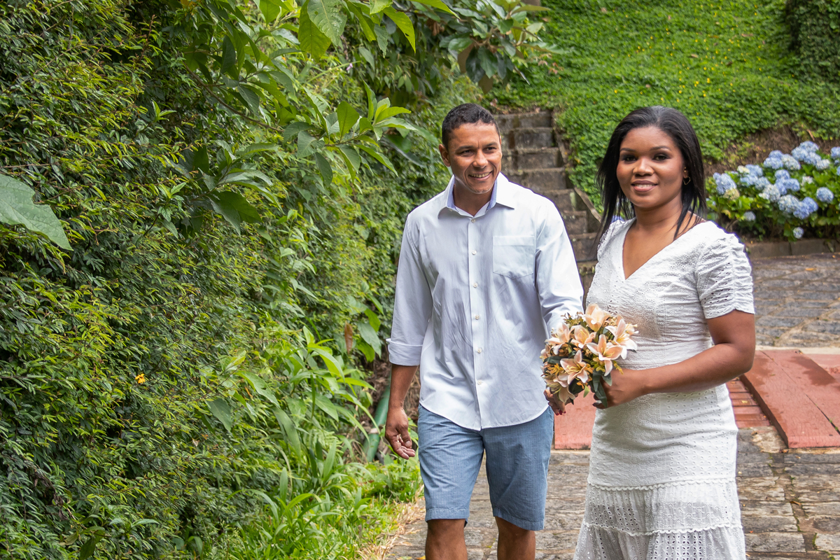 Noiva com buquê puxando a mão da noiva durante ensaio pré casamento   na serra de Teresópolis, noiva e noivo com roupas brancas em uma casa linda com muito verde e muitas flores e hortênsias azuis em Pàdua RJ.
