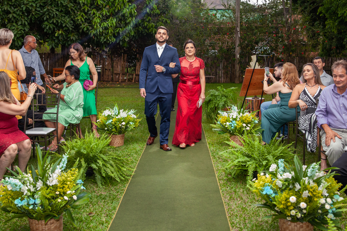 Noivo de terno azul entrando com sua mãe na cerimonia do seu casamento ao ar livre, sua mãe de vestido vermelho em Pádua interior do Rio de Janeiro.