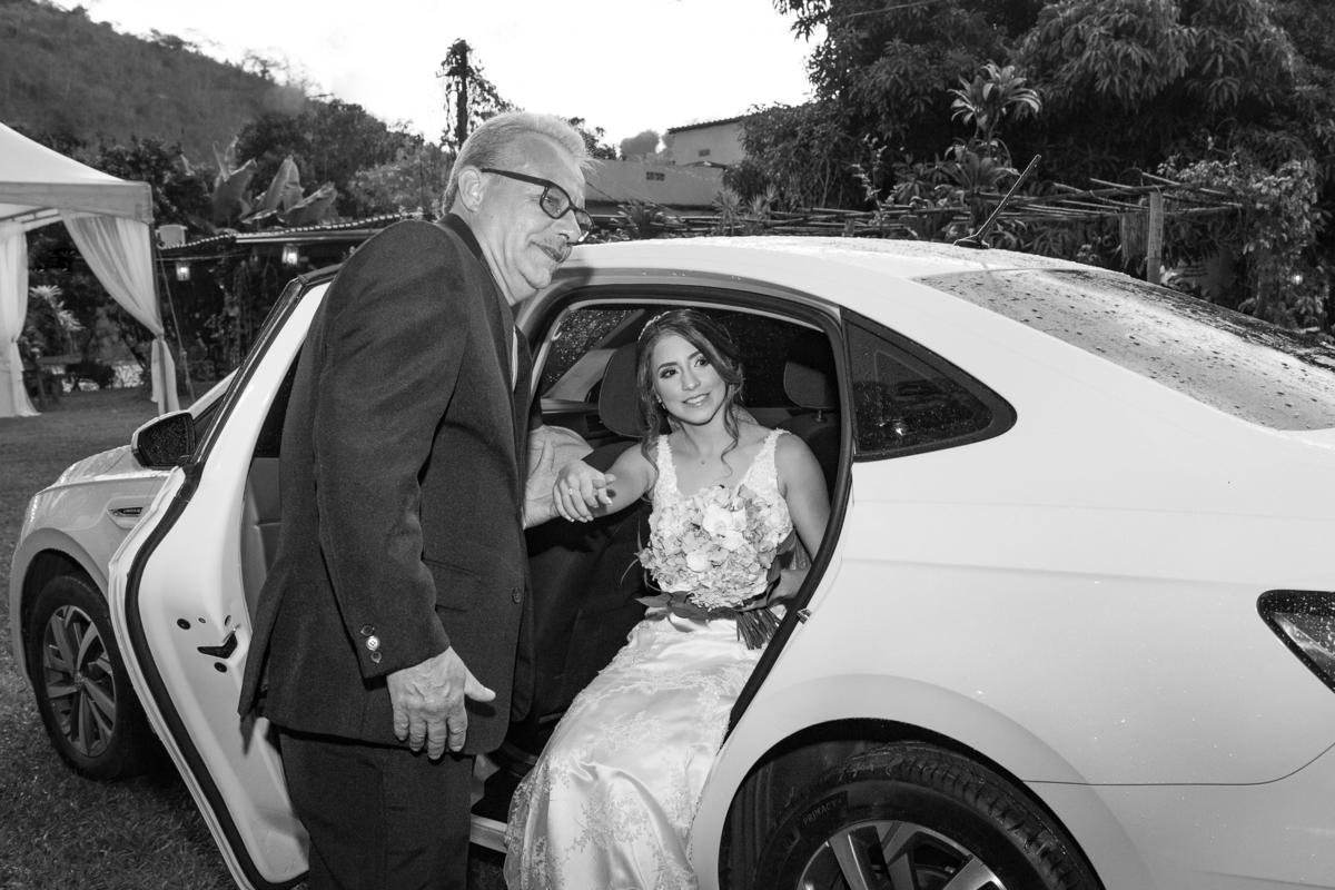 Noiva saindo do carro em foto preta e branca, o pai da noiva segurando sua mão minutos antes do casamento ao ar livre em Pádua interior do Rio de Janeiro.
