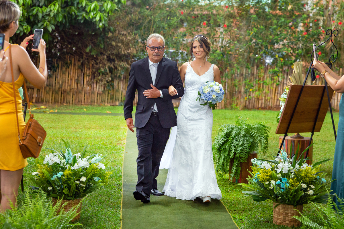 Noiva entrando em seu casamento com seu pai, noiva segurando um buque com flores naturais azuis e brancas em seu casamento ao ar livre em Pádua interior do Rio de Janeiro.