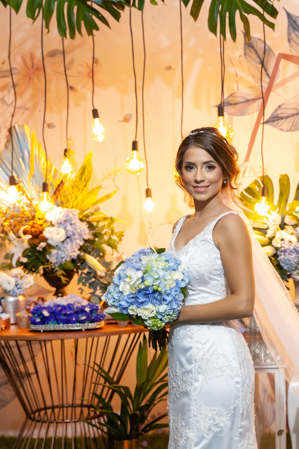 Noiva segurando o buque e sorrindo. decoração lâmpadas penduradas, segurando seu buque com flores azuis e brancas e a calda do vestido voando durante casamento em Santo Antônio de Pádua - Rj.