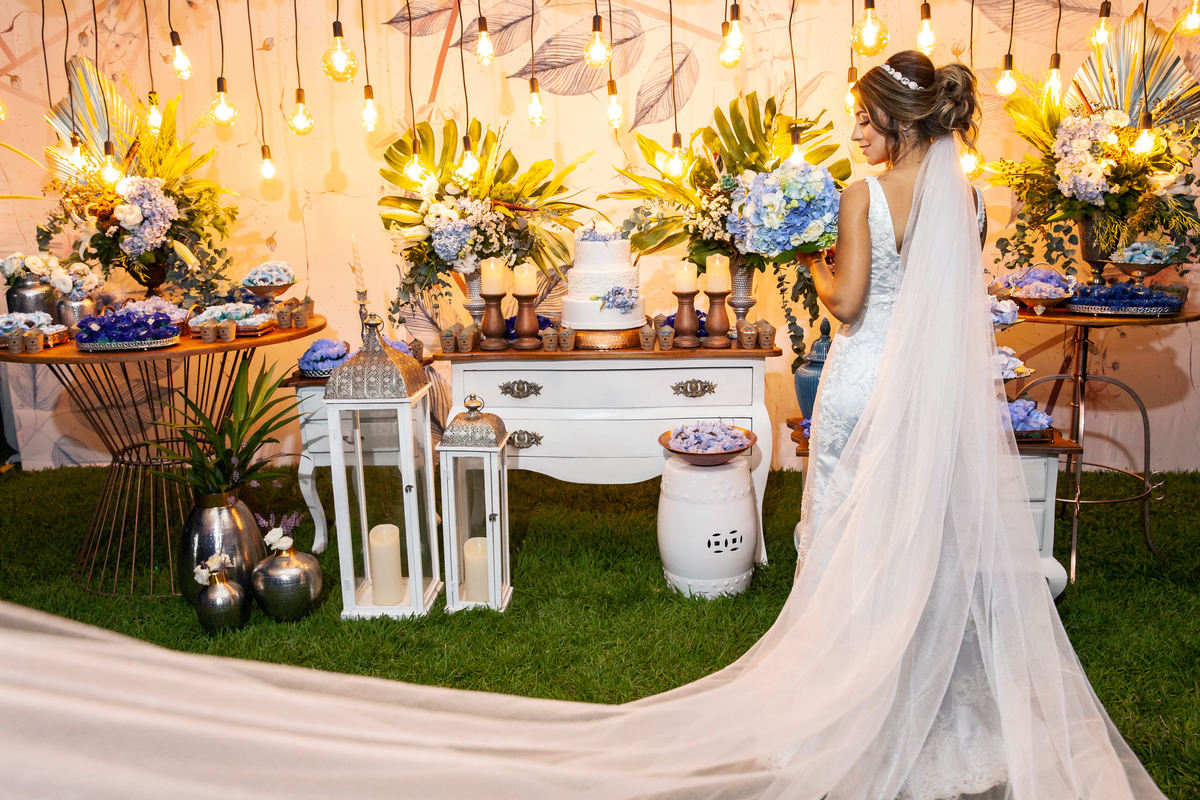 Noiva fazendo pose na decoração da sua festa com muitas lâmpadas penduradas, segurando seu buque com flores azuis e brancas e a calda do vestido voando durante casamento em Santo Antônio de Pádua - Rj.