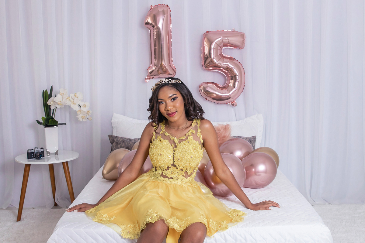 Ensaio 15 anos em estúdio, com um cenário de cama e número 15 bem grande atrás da cama. Bolas rosê gold. Maria linda vestida com vestido amarelo e coroa em Santo Antônio de Pádua no interior do Rio de Janeiro.