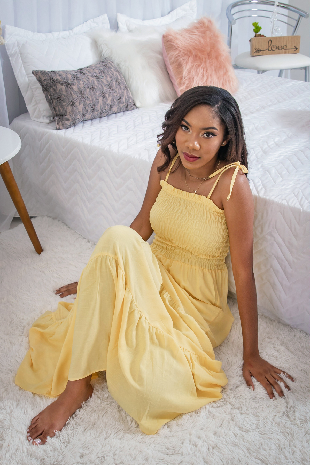 Ensaio 15 anos em estúdio, debutante com vestido amarelo encostada na cama sorrindo com a mão no queixo em Santo Antônio de Pádua no interior do Rio de Janeiro.