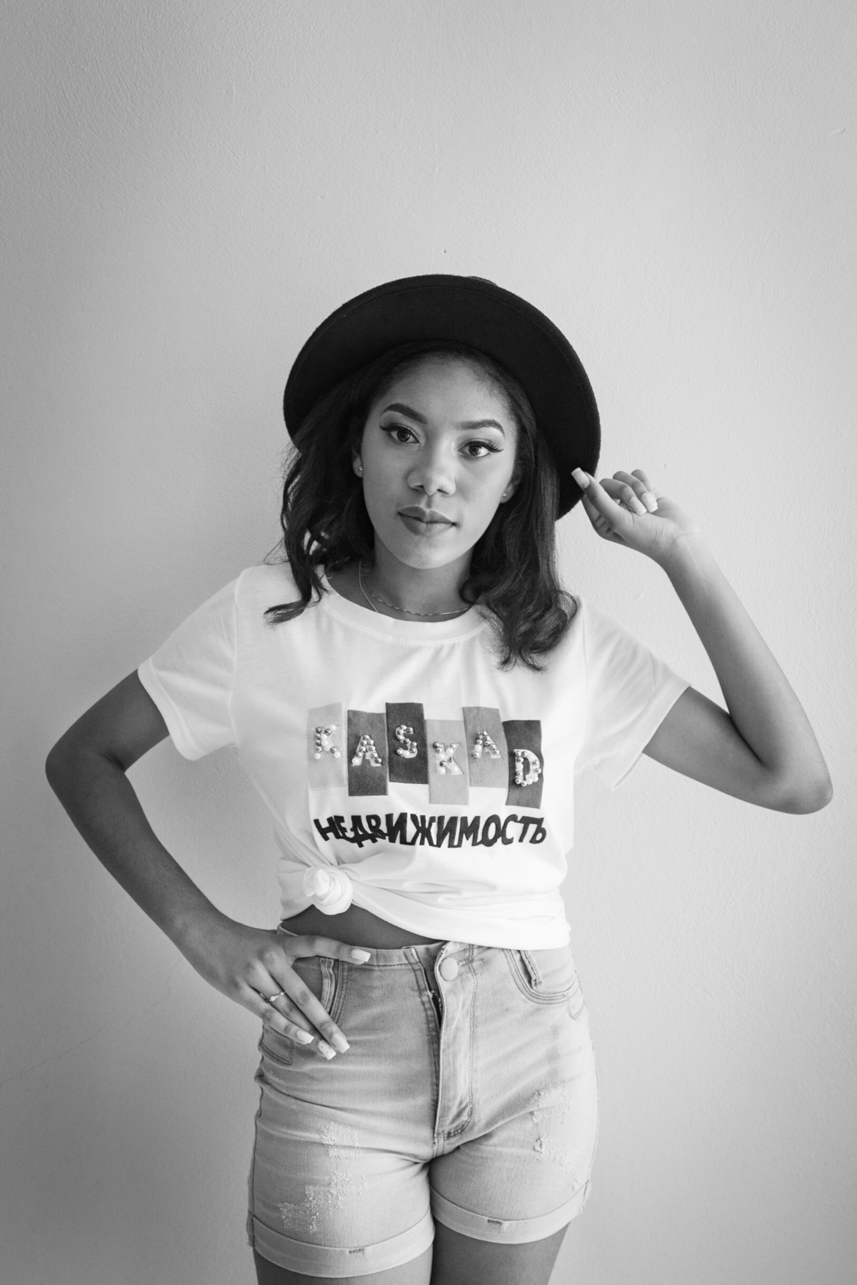 Ensaio 15 anos em estúdio, debutante short jeans e chapéu na cabeça encostada na parede sorrindo para suas fotos de 15 anos em Santo Antônio de Pádua no interior do Rio de Janeiro. Foto preta e branca.