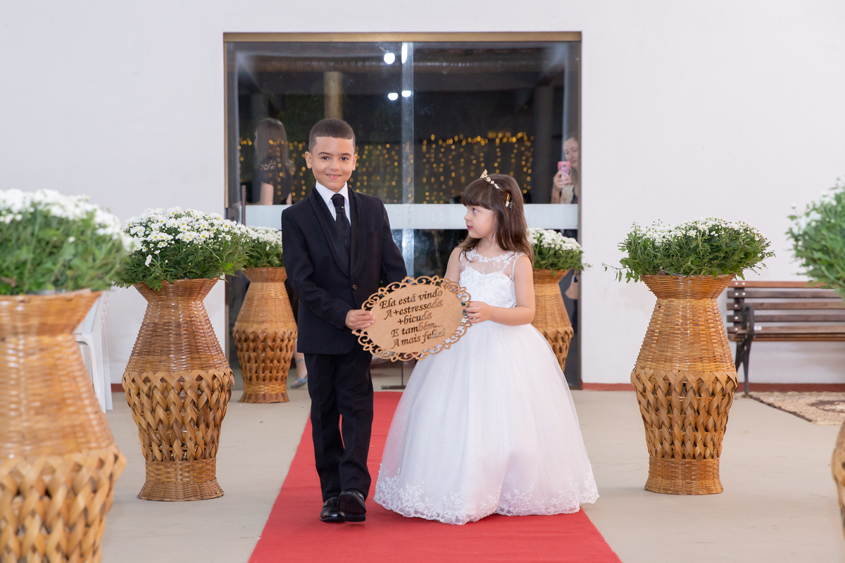 Dama e pajem entrando na cerimônia de casamento, segurando uma plaquinha. durante casamento durante a noite em um sitio em Cambuci no interior do Rio de Janeiro.