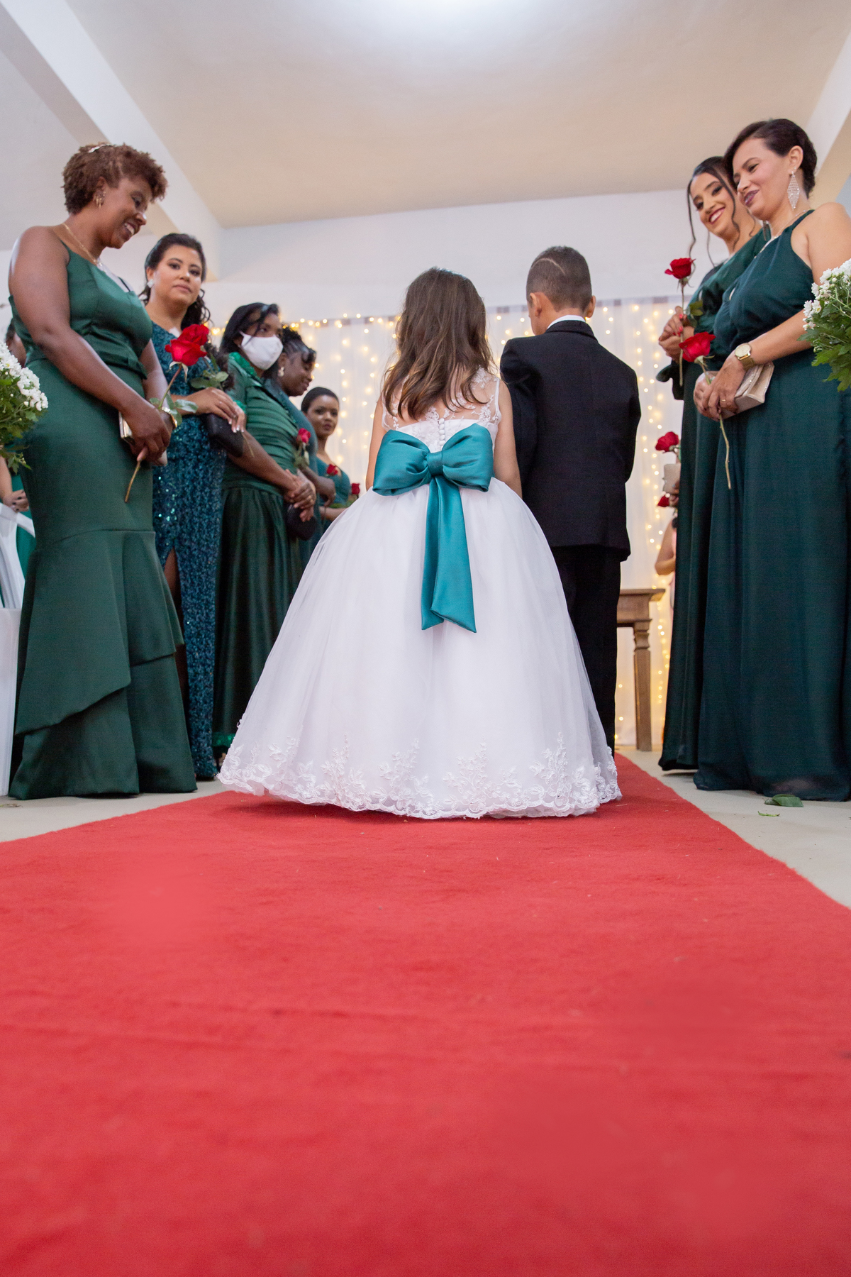 Dama e pajem entrando na cerimônia de casamento, segurando uma plaquinha. durante casamento durante a noite em um sitio em Cambuci no interior do Rio de Janeiro. Madrinhas com vestido verde olhando e segurando uma rosa vermelha nas mãos.