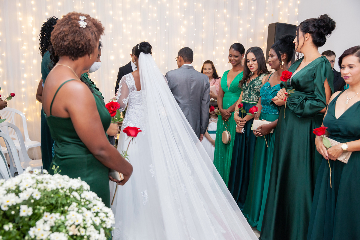 Noiva com vestido de renda e véu lindo e noivo com terno cinza durante casamento durante a noite em um sitio em Cambuci no interior do Rio de Janeiro. Madrinhas de vestido verde escuro olhando para os noivos com rosas vermelhas na mão.