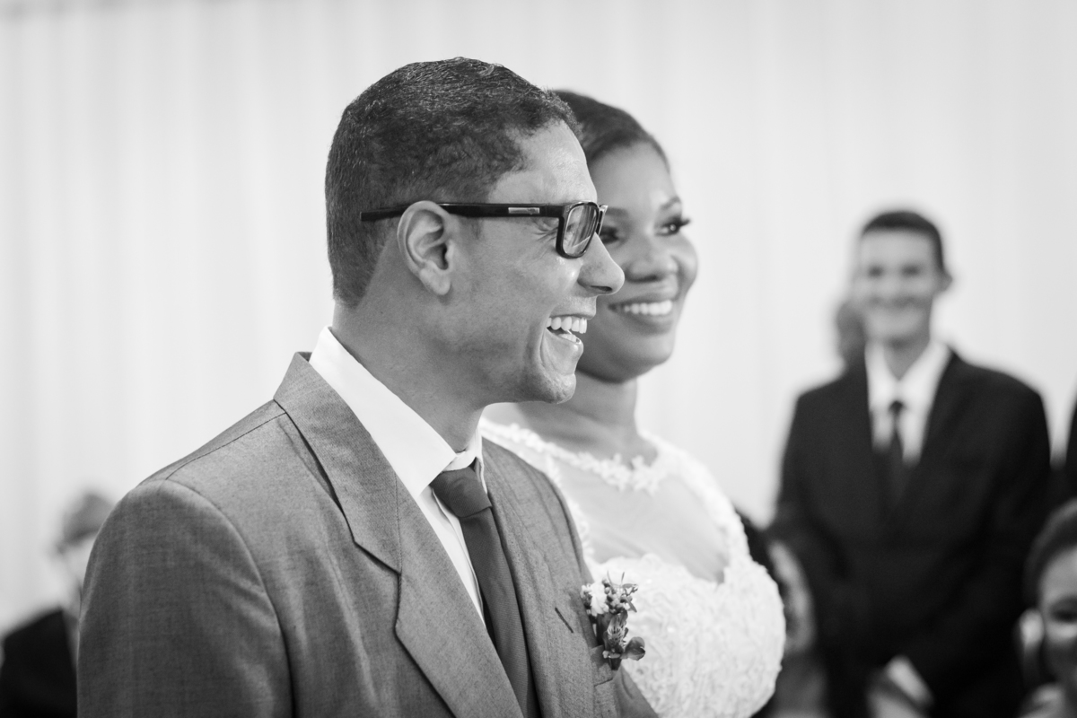 Noiva com vestido de renda e véu lindo e noivo com terno cinza se olhando e sorrindo durante cerimônia casamento durante a noite em um sitio em Cambuci no interior do Rio de Janeiro. Foto preta e branca.