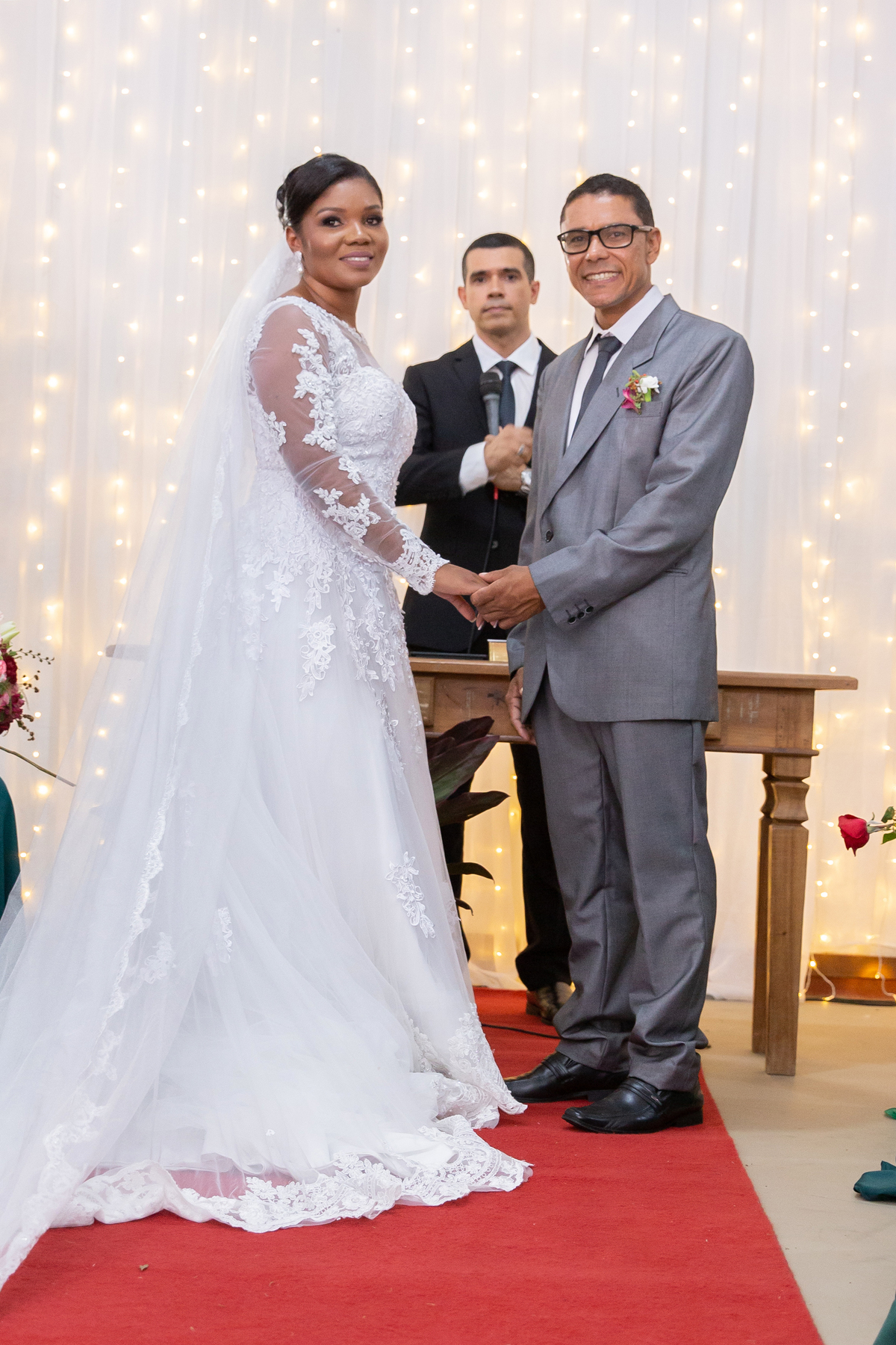 Noiva com vestido de renda e véu lindo e noivo com terno cinza se olhando e sorrindo durante cerimônia casamento durante a noite em um sitio em Cambuci no interior do Rio de Janeiro. 