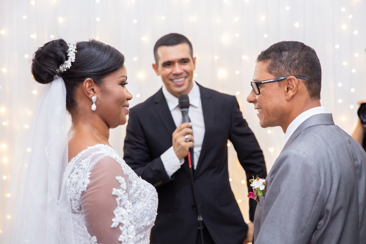 Noiva com vestido de renda e véu lindo e noivo com terno cinza se olhando e sorrindo durante cerimônia casamento durante a noite em um sitio em Cambuci no interior do Rio de Janeiro. 