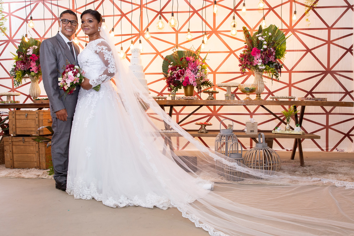 Noiva com vestido de renda e véu lindo e noivo com terno cinza. Noivos fazendo fotos na mesa do bolo com decoração de lâmpadas penduradas em  casamento durante a noite em um sitio em Cambuci no interior do Rio de Janeiro.