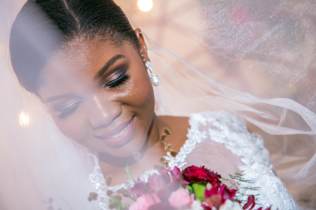 Noiva com vestido de renda e véu lindo fazendo fotos com seu buquê com flores em tons de vermelho durante cerimônia evangélica em casamento durante a noite em um sitio em Cambuci no interior do Rio de Janeiro.