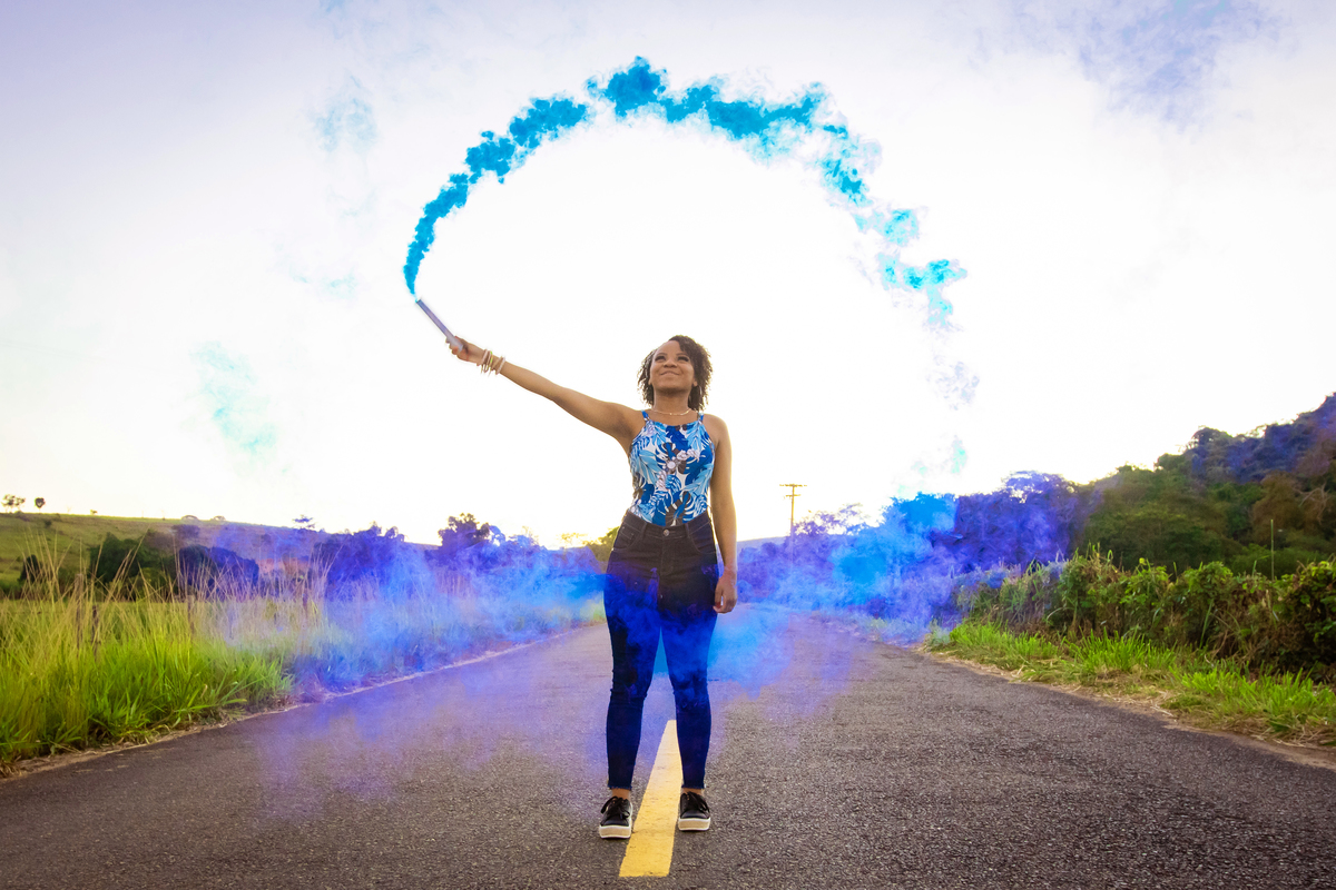 Aniversariante linda de 15 anos, com cabelo cacheado em um dia lindo com muito verde ao por do sol lindo no meio do asfalto em seu ensaio de 15 Anos. No interior do Rio de Janeiro. Com fumaça azul para a foto.