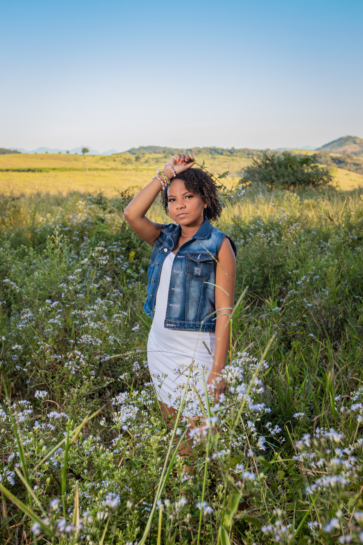 Aniversariante linda com jaqueta jeans em um campo florido com um céu azul perfeito no parque municipal de Miracema em seu ensaio de 15 Anos. No interior do Rio de Janeiro. 
