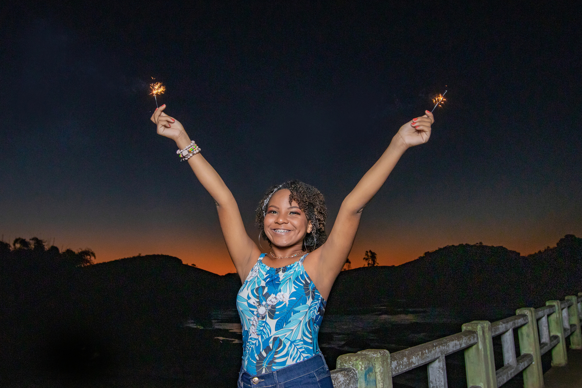 Aniversariante linda de 15 anos, com cabelo cacheado, segurando uma vela  em um dia lindo ao por do sol perfeito, com céu laranja em seu ensaio de 15 Anos. No interior do Rio de Janeiro.