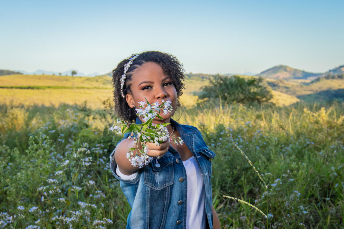 Aniversariante linda com jaqueta jeans em um campo florido com um céu azul perfeito no parque municipal de Miracema em seu ensaio de 15 Anos. No interior do Rio de Janeiro.