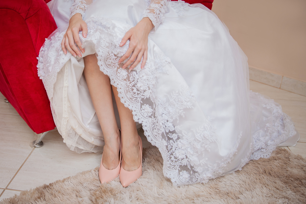 Noiva colocando o sapato com seu vestido lindo com flores em 3D sentada em sofá vermelho vermelho durante making of em salão de beleza em Pirapetinga para casamento em Santo Antônio de Pádua interior do Rio de Janeiro.