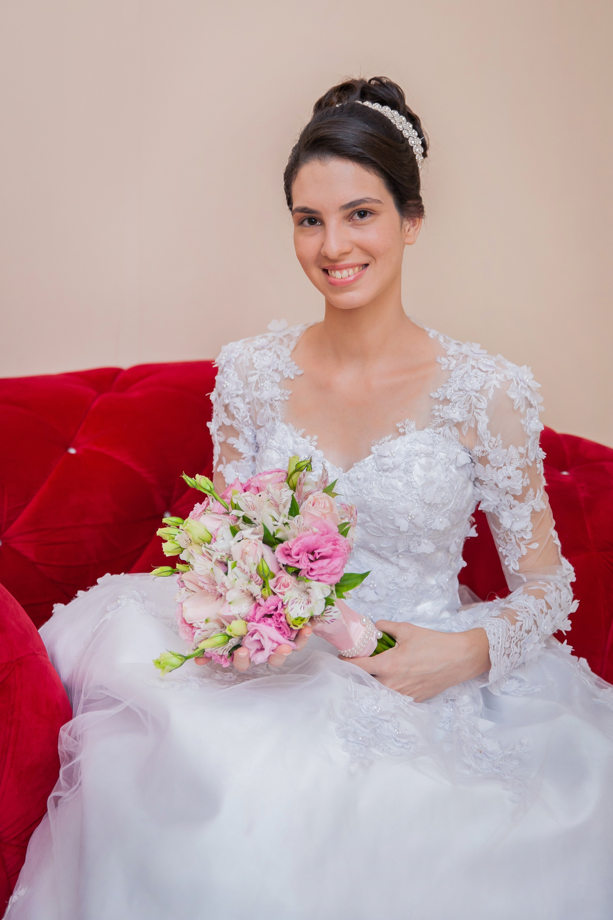 Noiva segurando o buquê com flores em tons de rosas com seu vestido lindo com flores em 3D sentada em sofá vermelho vermelho durante making of em salão de beleza em Pirapetinga para casamento em Santo Antônio de Pádua interior do Rio de Janeiro.