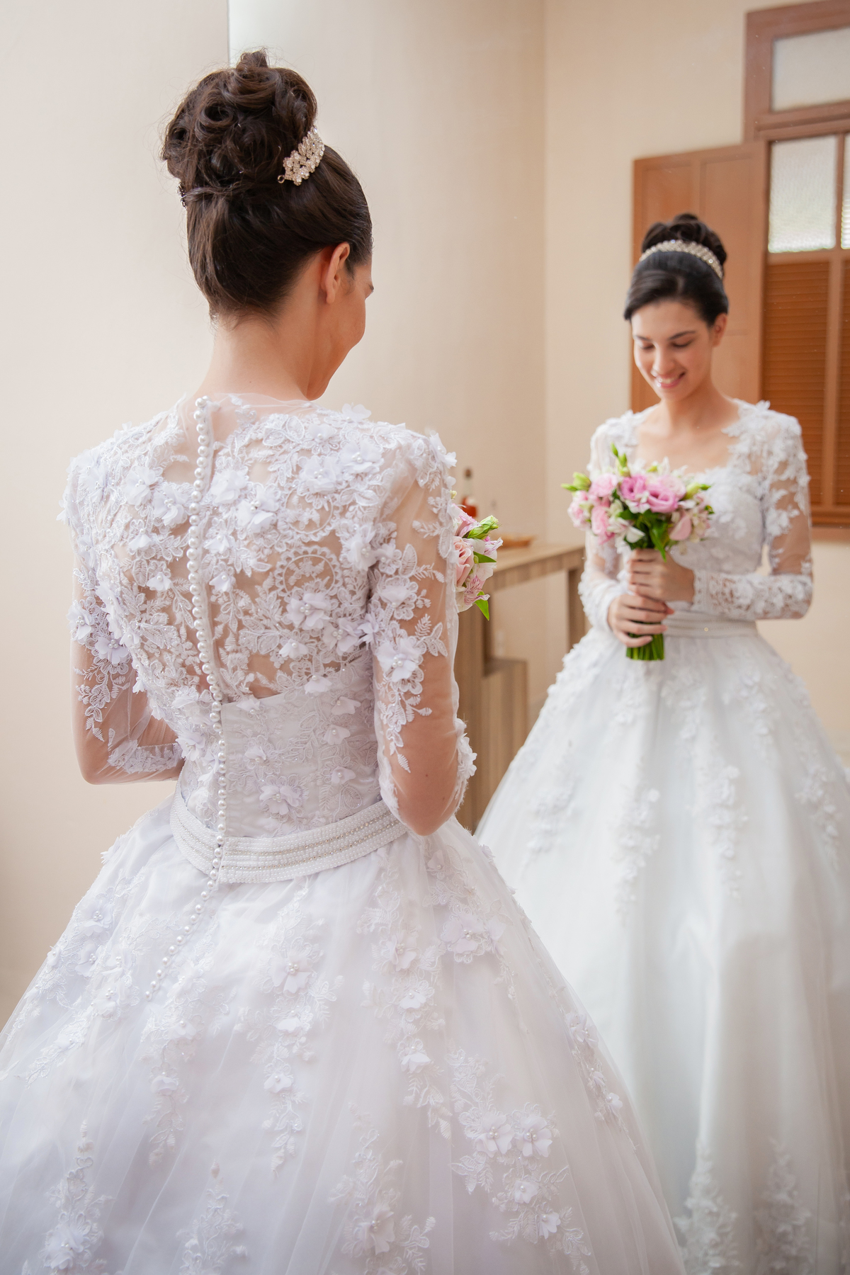 Noiva segurando o buquê com flores em tons de rosas com seu vestido lindo com flores em 3D de frente para o espelho e sorrindo durante making of em salão de beleza em Pirapetinga para casamento em Santo Antônio de Pádua interior do Rio de Janeiro.
