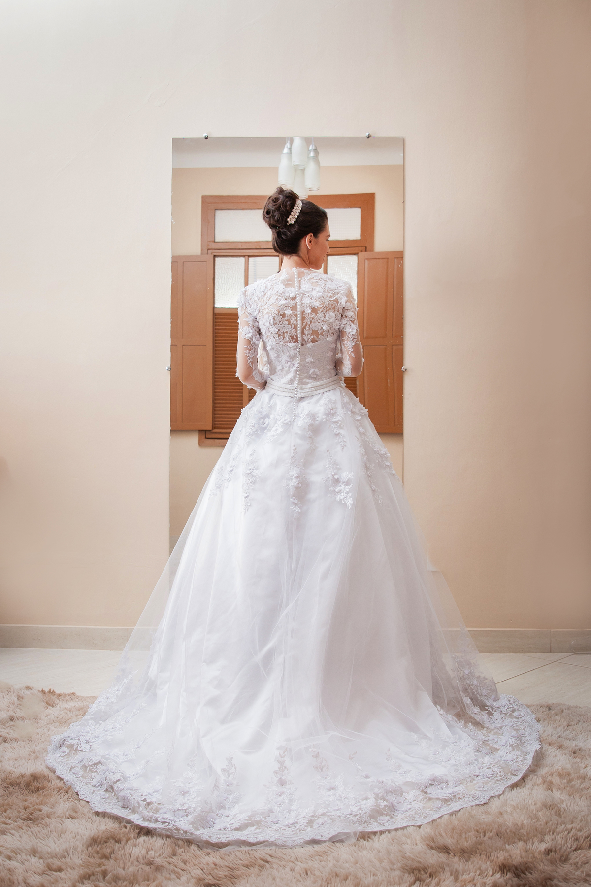 Noiva segurando o buquê com flores em tons de rosas com seu vestido lindo com flores em 3D de frente para o espelho e sorrindo durante making of em salão de beleza em Pirapetinga para casamento em Santo Antônio de Pádua interior do Rio de Janeiro.