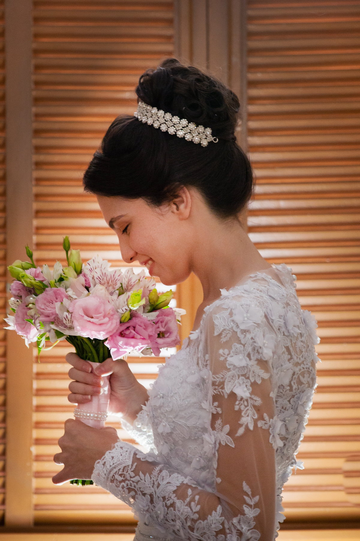 Noiva cheirando seu buquê com flores em tons de rosas com seu vestido lindo com flores em 3D de frente perto da janela durante making of em salão de beleza em Pirapetinga para casamento em Santo Antônio de Pádua interior do Rio de Janeiro.
